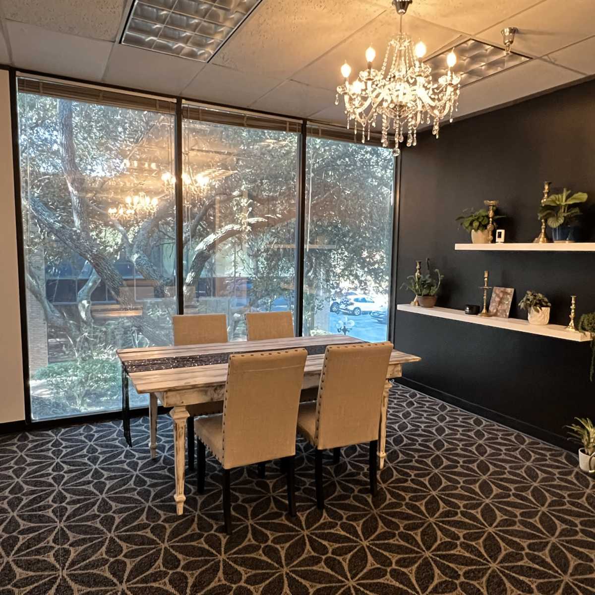A modern meeting room with a wooden table surrounded by four beige chairs, large windows providing natural light, and a chandelier hanging from the ceiling.