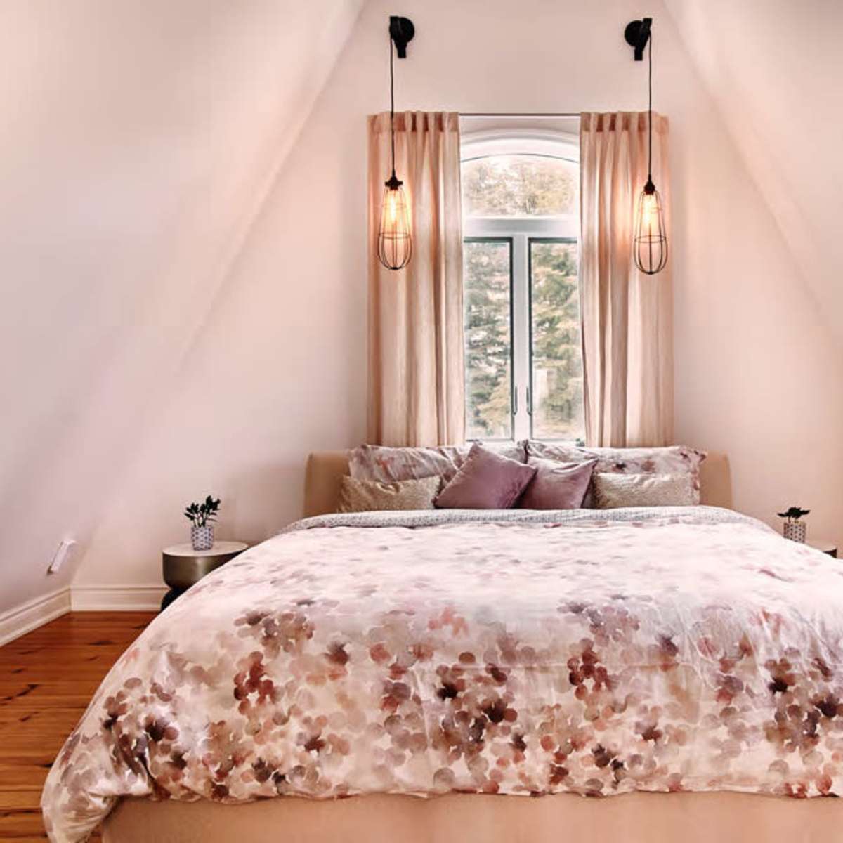A well-lit bedroom features an angled ceiling, a large bed with a floral patterned duvet, and two pendant lights above.