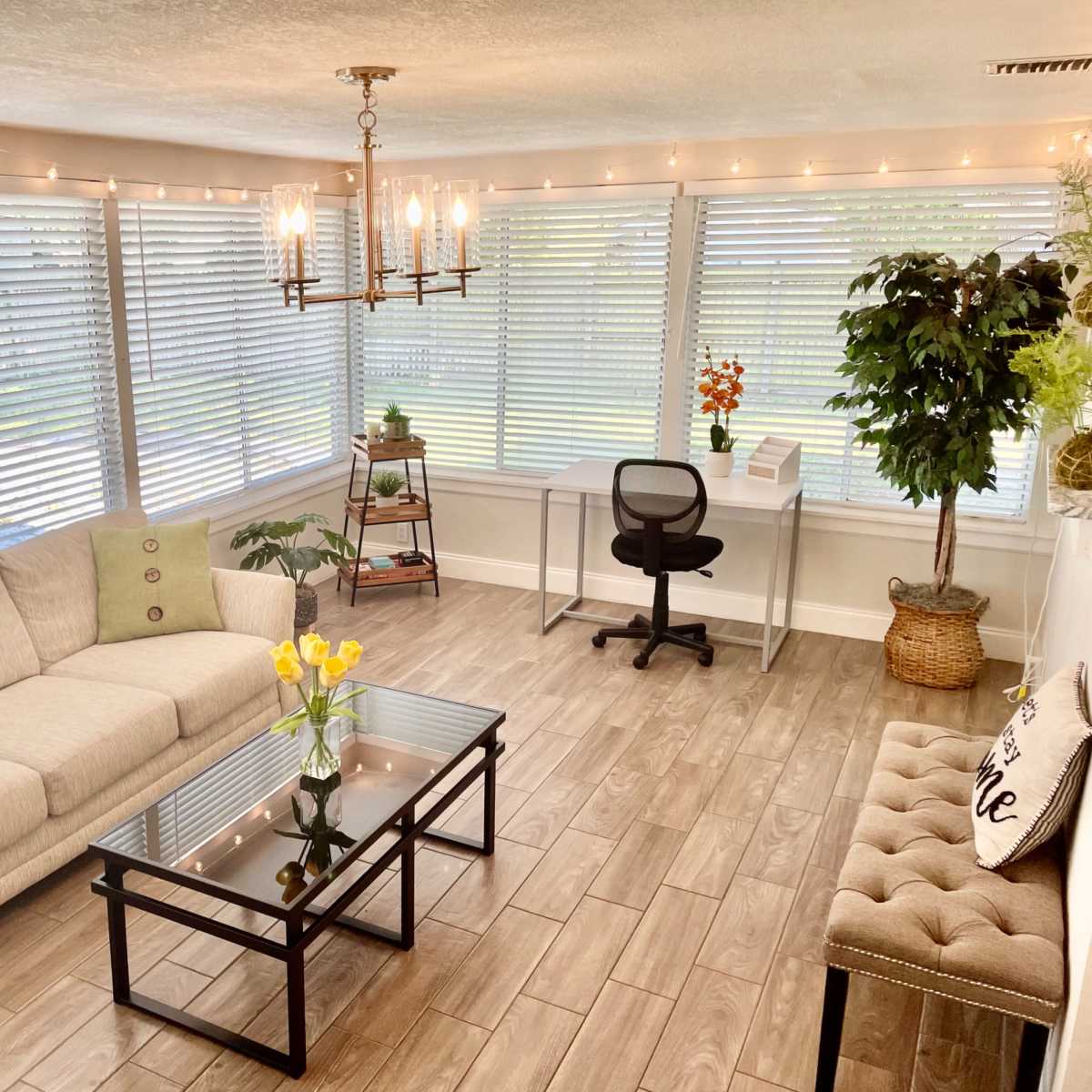 A well-lit room with large windows covered by blinds, featuring a light-colored sofa, a glass coffee table with a vase of yellow flowers, a black office chair, and a small desk with a decorative plant in the corner.