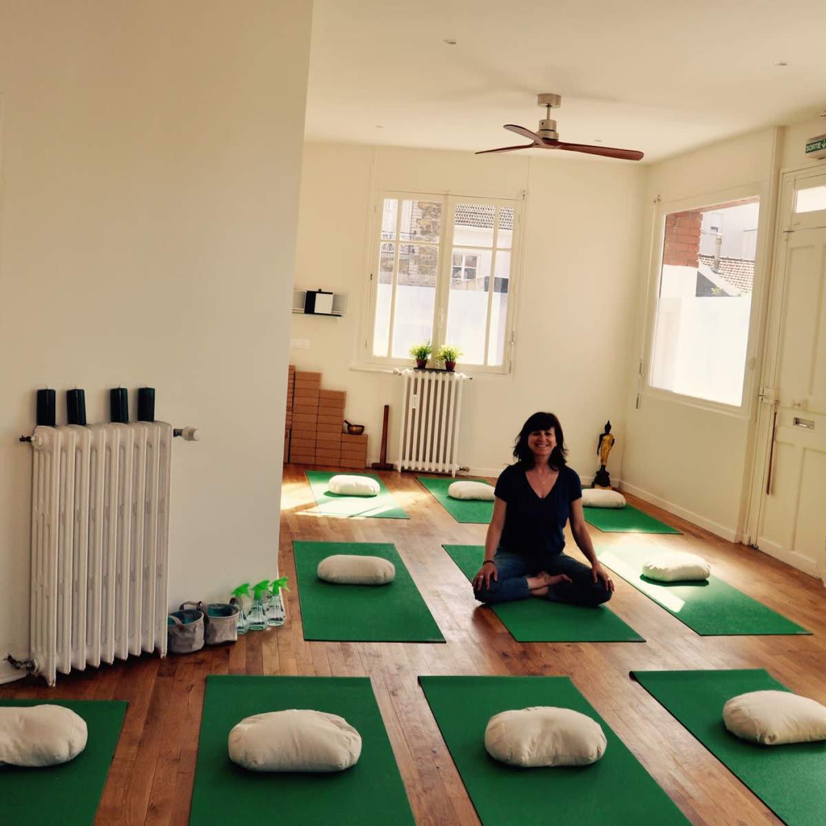 A person sits cross-legged on a green yoga mat in a minimalist studio with additional mats and cushions arranged around her.