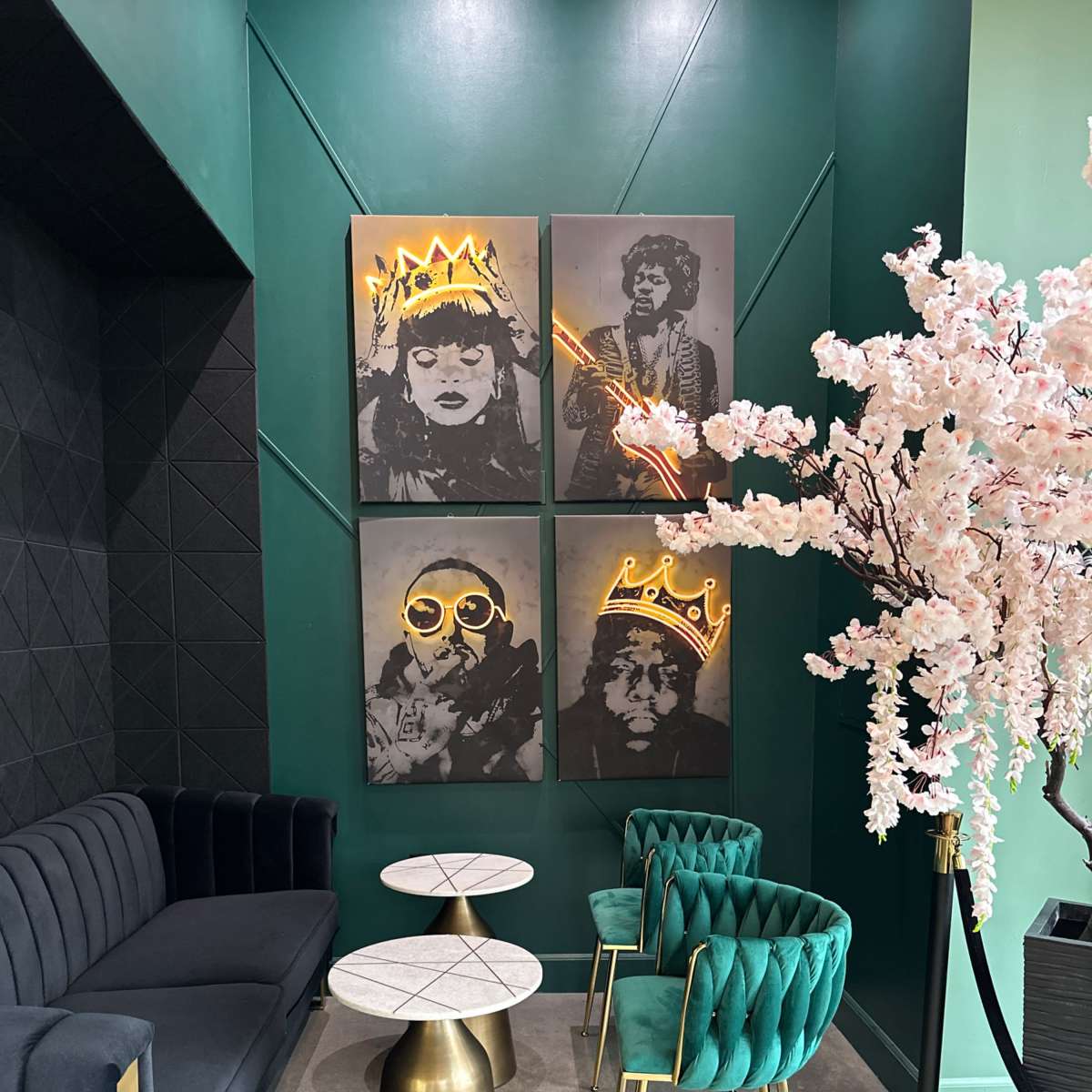 A corner of a stylish café with two marble-top tables, green velvet chairs, a black sofa, and four framed portraits with neon accents on the walls, alongside a pink cherry blossom tree.