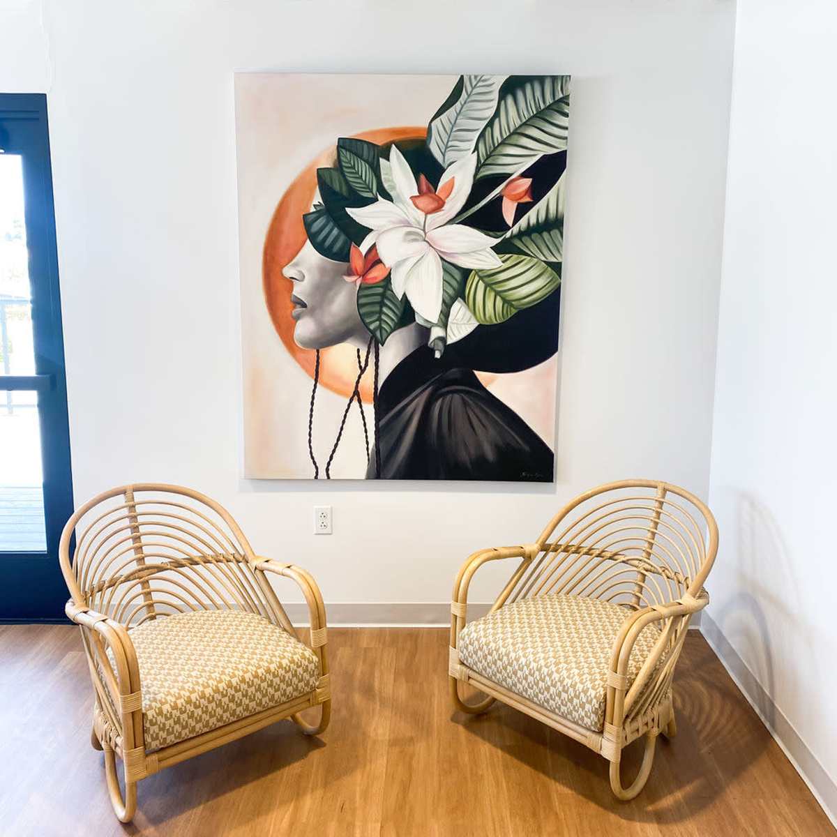 A pair of rattan chairs facing a large painting of a woman with floral elements on her head, positioned in a well-lit room with wooden flooring.