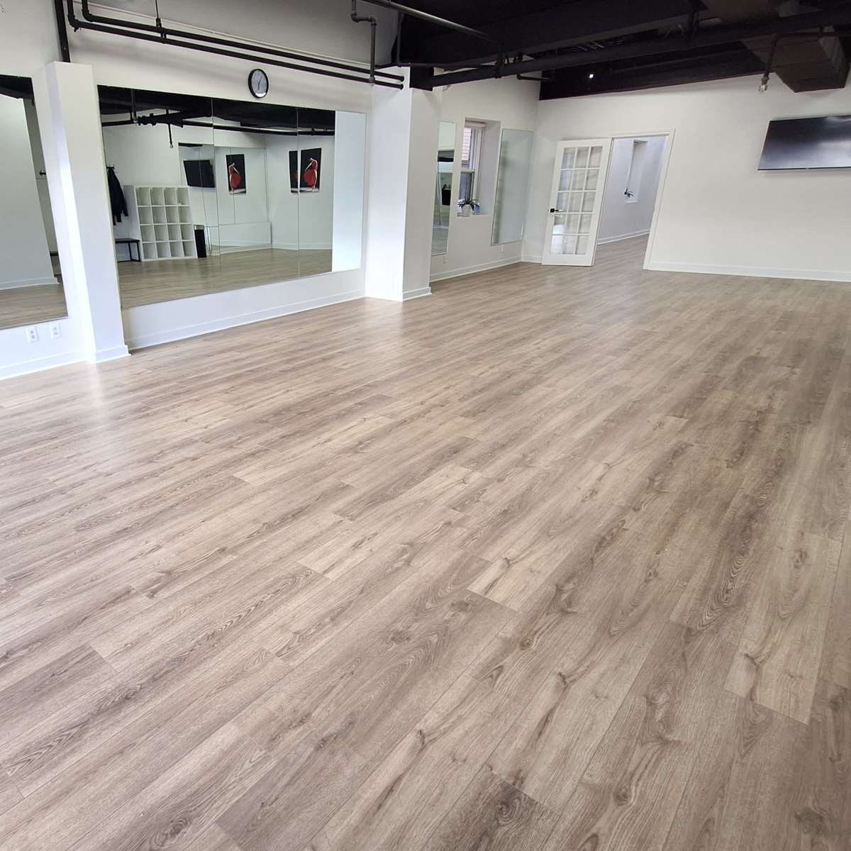 An empty, well-lit studio space with wooden flooring, mirrors on one wall, and an adjoining room visible through an open door.