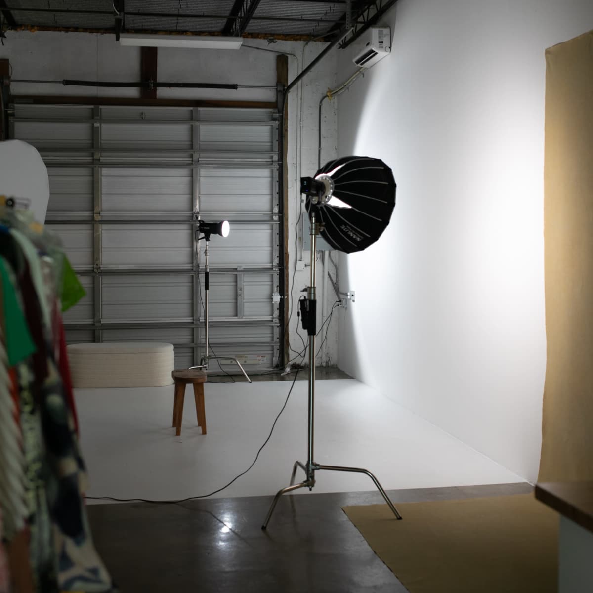 A photography studio features two light sources and a plain white wall, with clothing hanging on a rack in the foreground.
