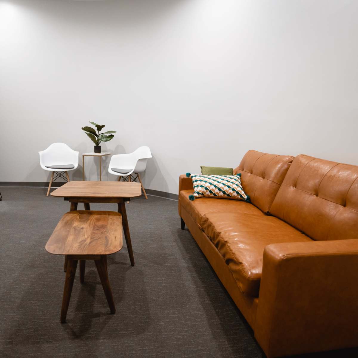 A cozy waiting area featuring a brown leather couch, two white chairs, a small table, and a large mirror against a plain wall.