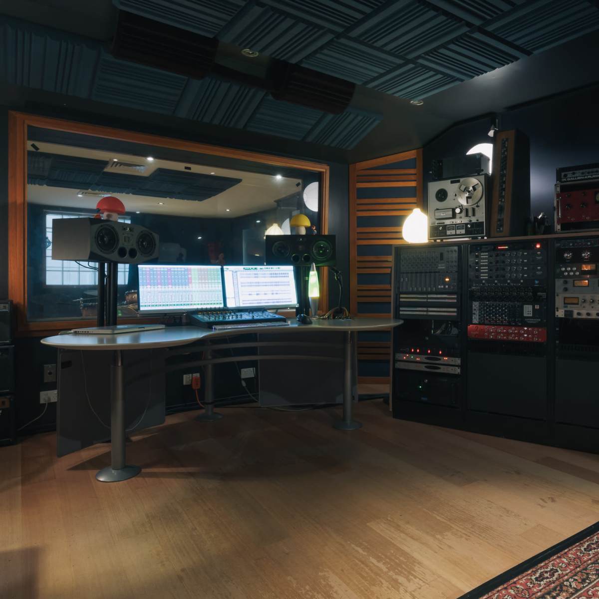 A music recording studio features a mixing desk surrounded by audio equipment, speakers, and a decorative lamp.