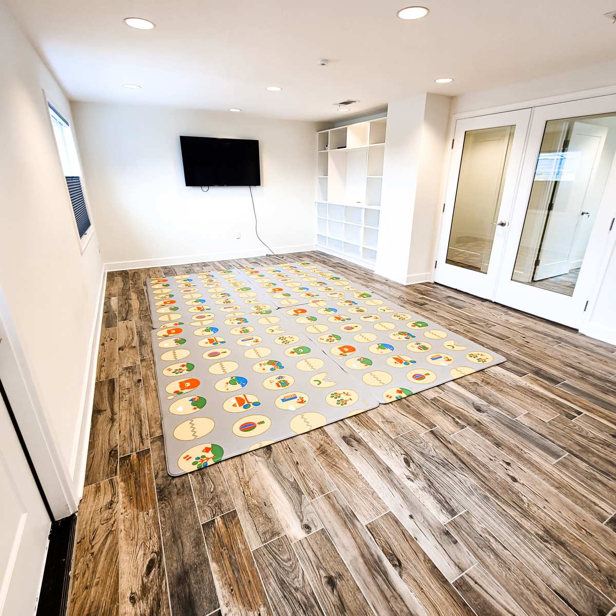 The room features a large area rug with a playful design on a wooden floor, accompanied by a wall-mounted TV and built-in shelving.