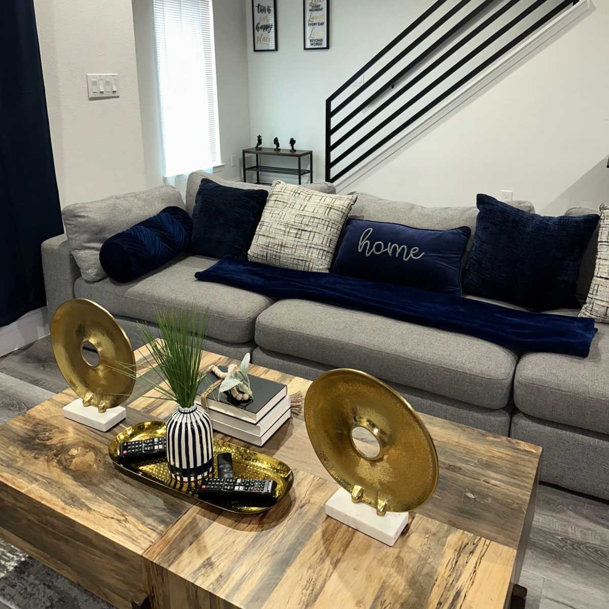 A modern living room featuring a gray sofa adorned with decorative pillows, a wooden coffee table with a decorative tray, and a staircase in the background.