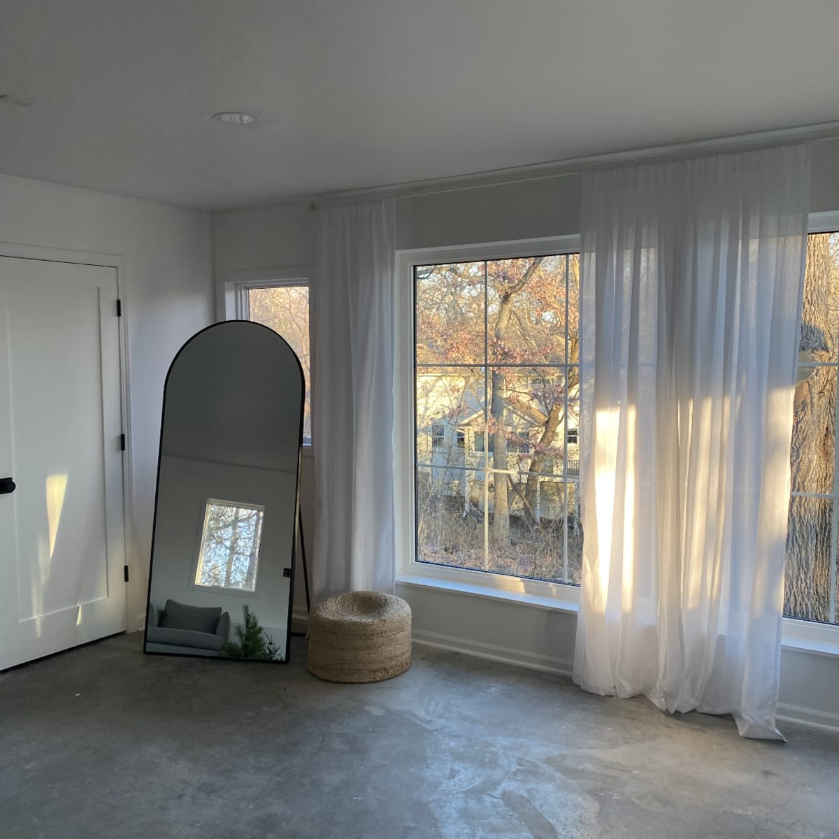 A minimalist room features a tall mirror, large windows with sheer curtains, and a round woven basket.