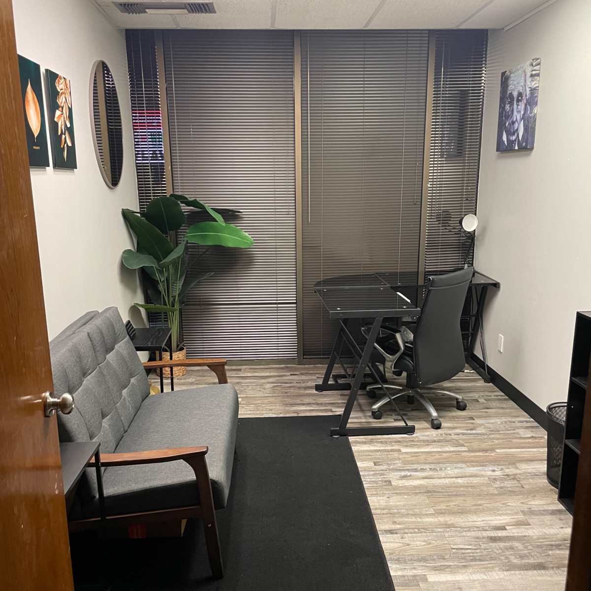 A small office space with a gray couch, a desk with a chair, and a large plant near a window with blinds.