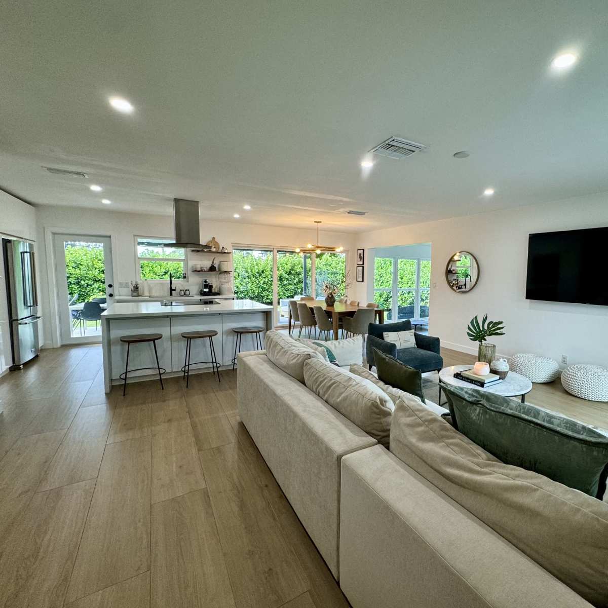 A modern, open-concept living room with a sofa facing a wall-mounted TV and a dining area in the background, surrounded by large windows.