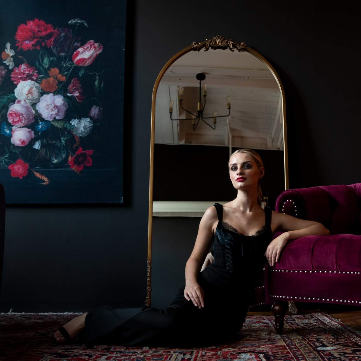 A woman in a black dress poses next to a burgundy velvet sofa, reflecting in a round mirror against a dark wall with a floral painting nearby.
