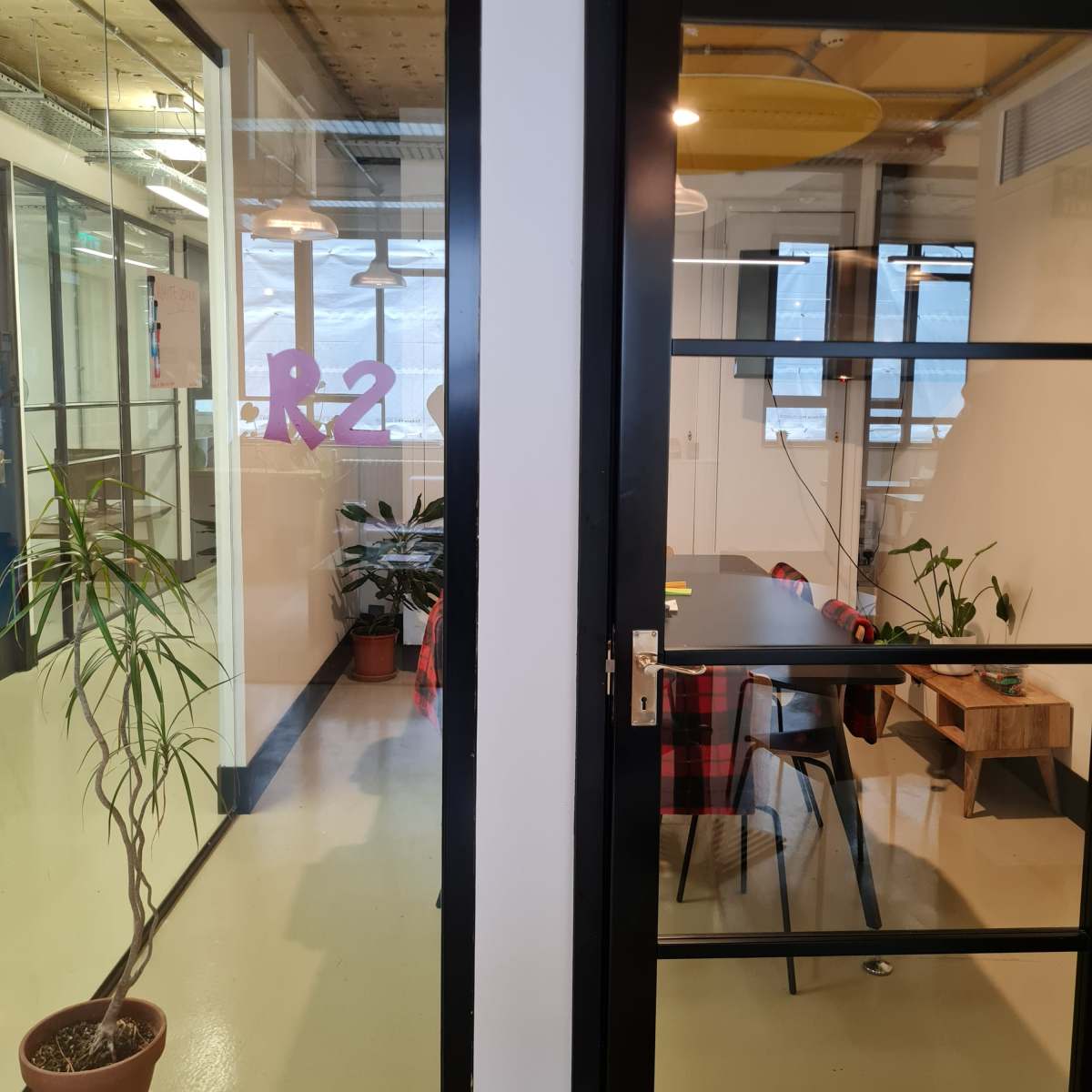 Two adjacent glass-walled office spaces, one labeled "R2," with plants and a table visible in each room.