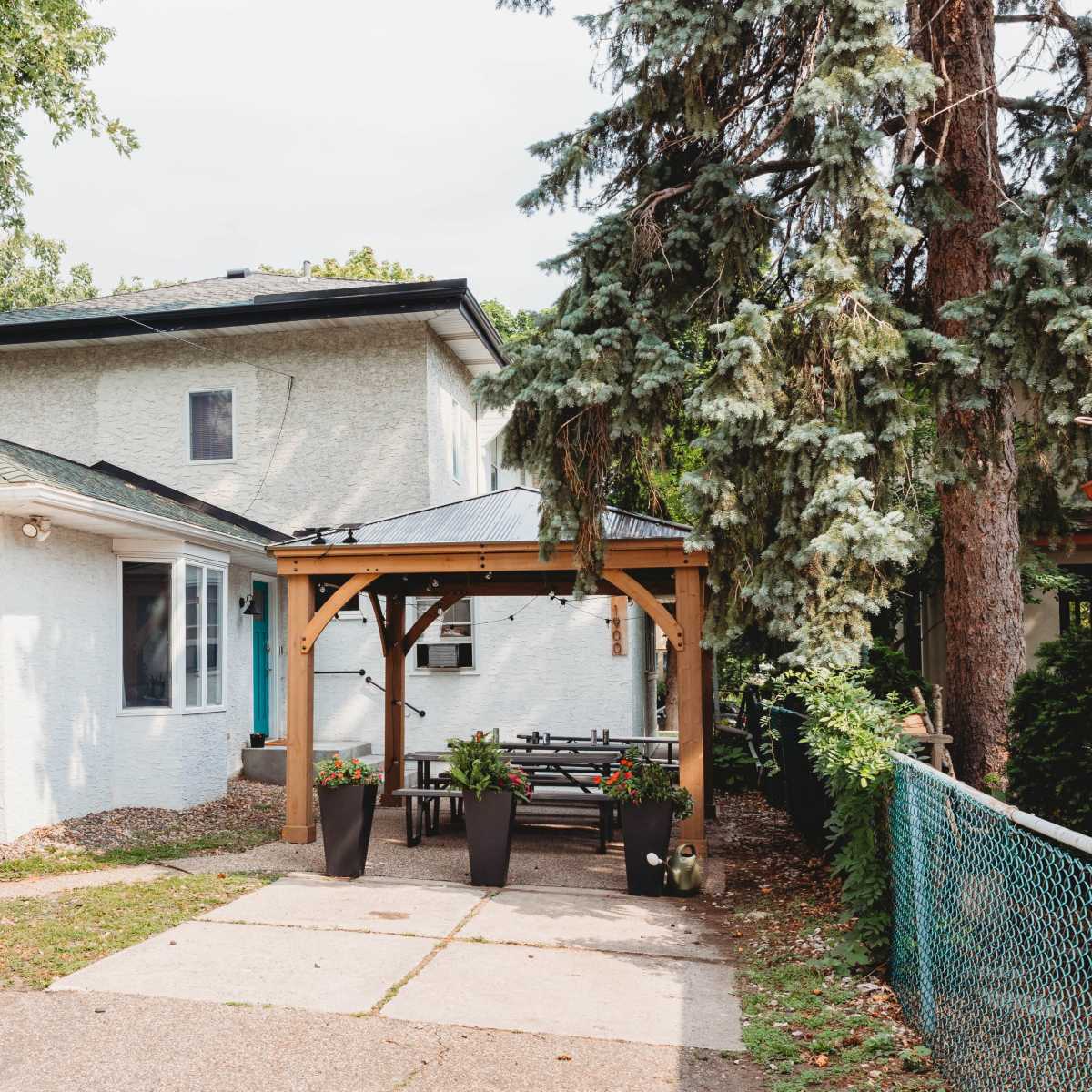 A two-story, light-colored house is shown with a paved driveway, a covered outdoor seating area, and trees alongside the property.