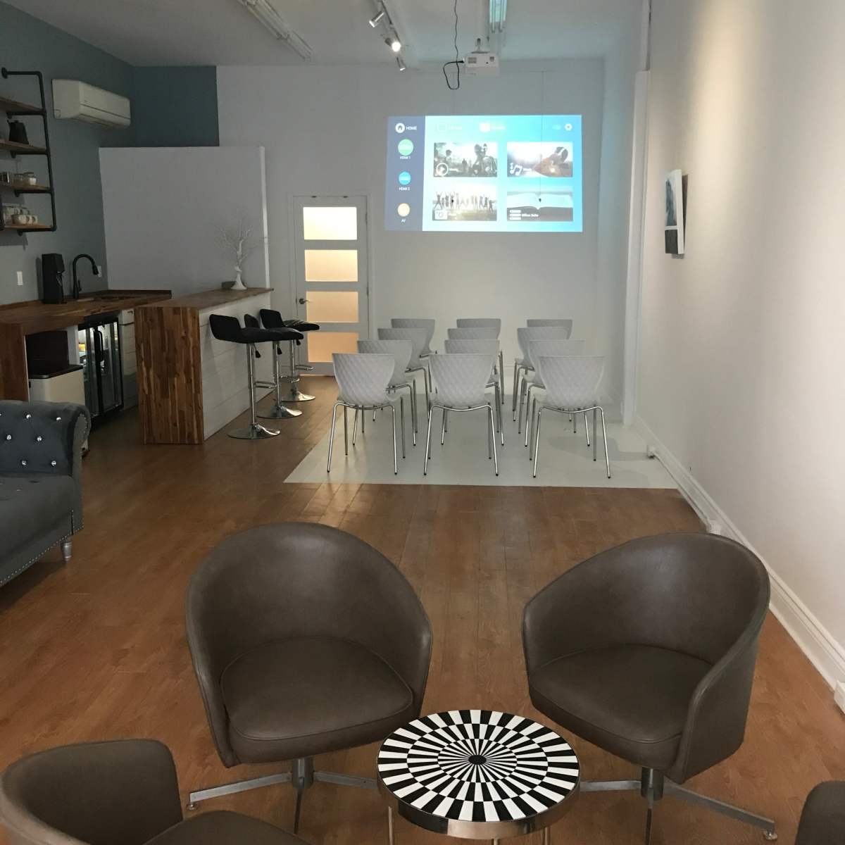 A modern meeting space featuring a row of white chairs facing a projected screen, with cozy seating and a wooden bar area in the background.