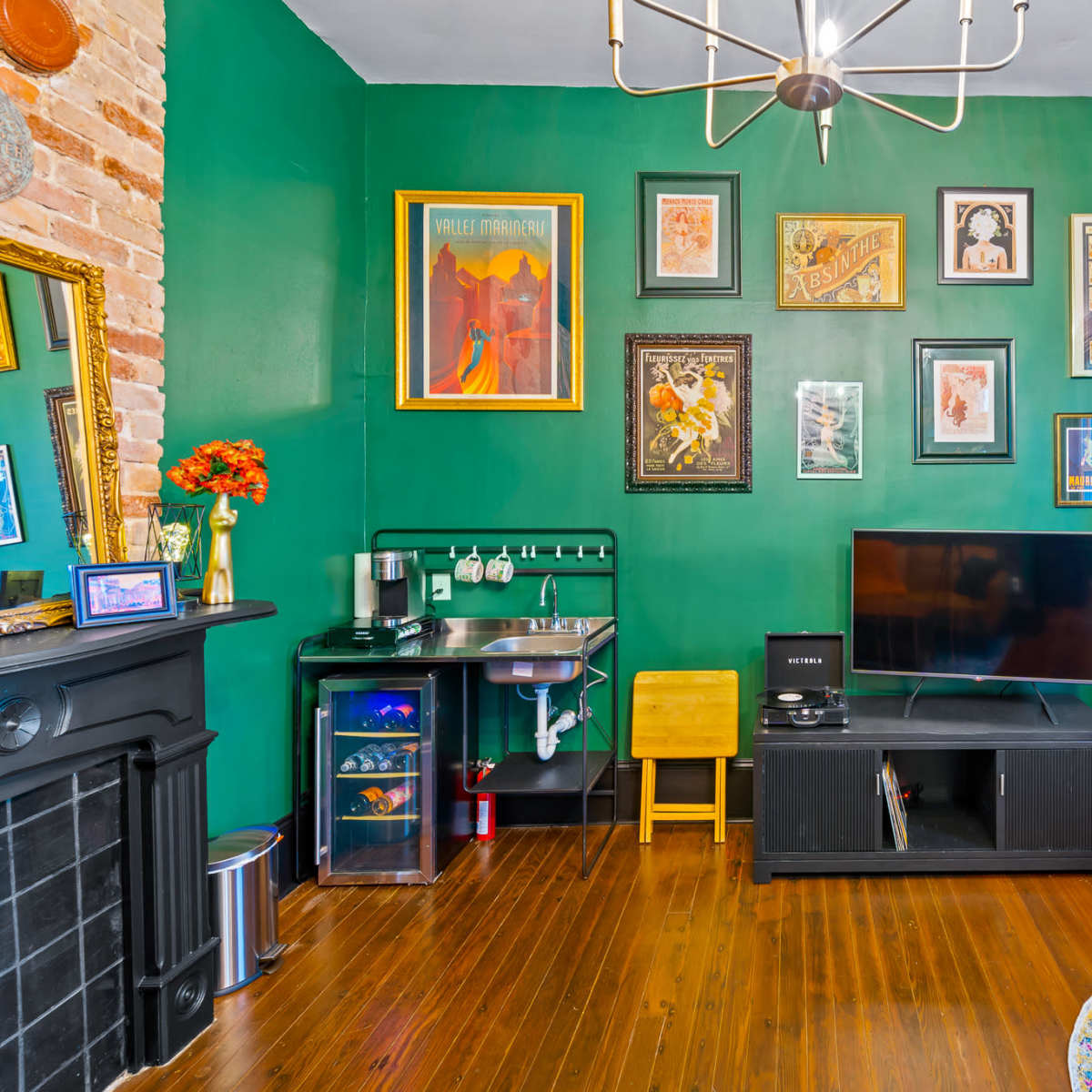 The scene features a room with green walls adorned with various framed artworks, a black TV stand with a television, a small kitchenette area, and a blue chair beside a potted plant.