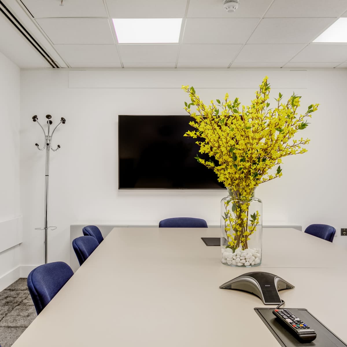 A meeting room features a large table surrounded by blue chairs, a wall-mounted television, and a vase of yellow flowers on the table.