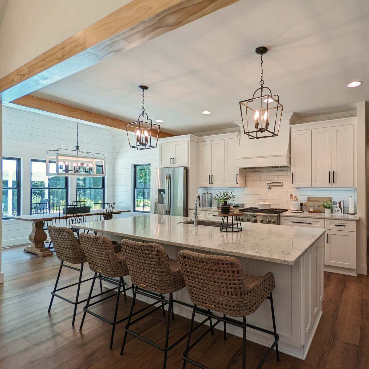 A modern kitchen with white cabinetry, a large island with bar stools, and a dining area featuring a wooden table and large windows.