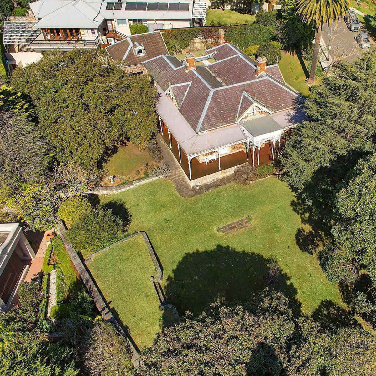 An aerial view of a house surrounded by greenery and trees, featuring a large lawn and a pathway.