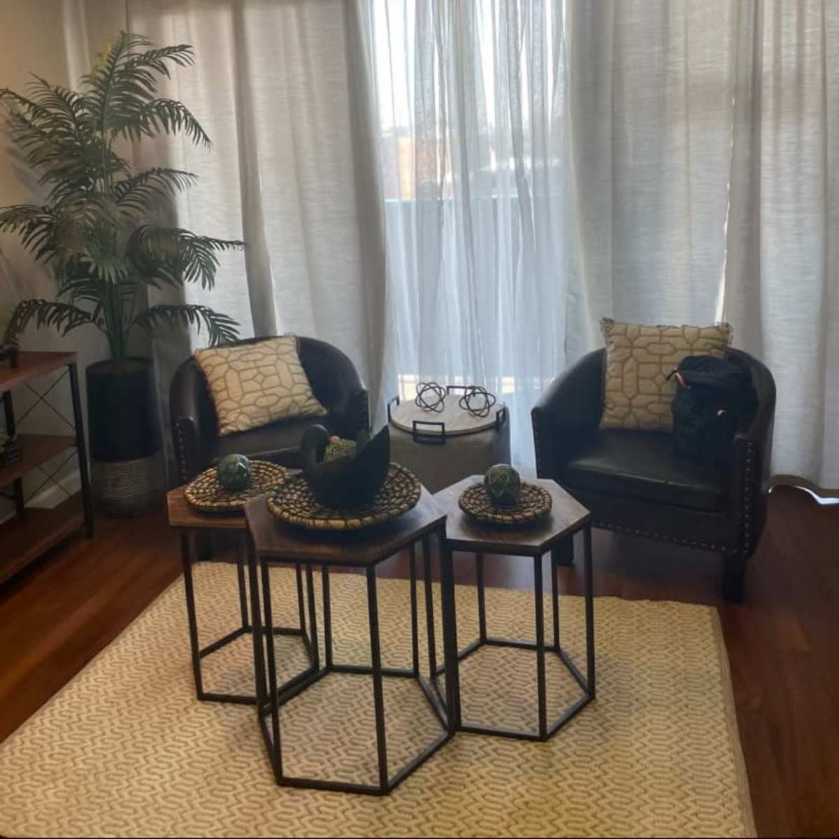 A well-lit office space featuring two black leather chairs with patterned cushions, a set of three hexagonal tables, and a large window dressed with sheer white curtains.