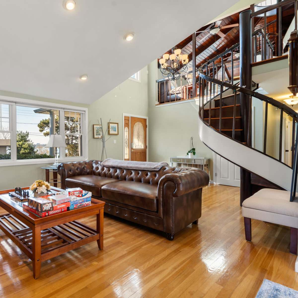 A spacious living room features a brown leather sofa, a wooden coffee table with board games, and a winding staircase leading to an upper level.