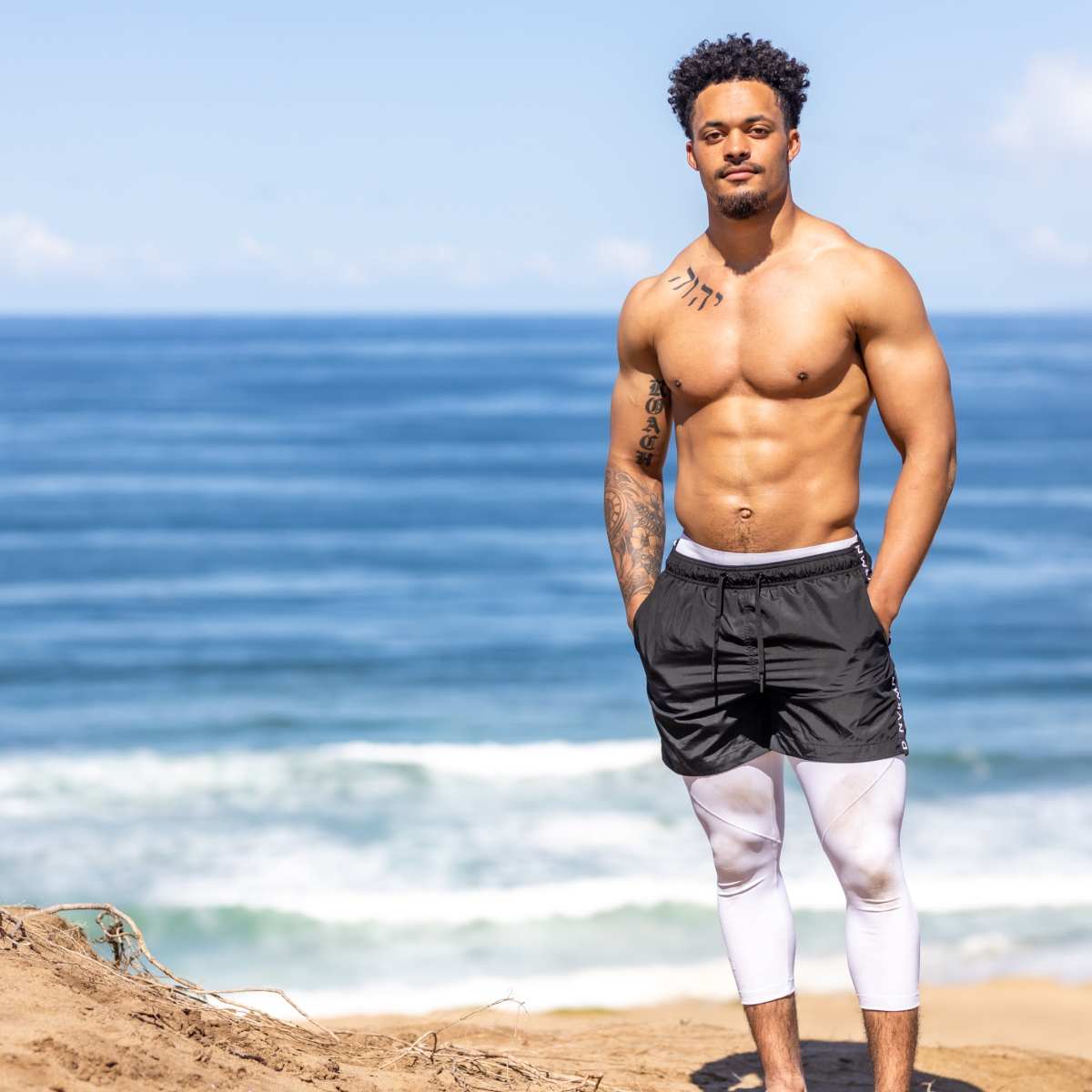 A shirtless man with tattoos stands on a sandy shoreline overlooking the ocean.