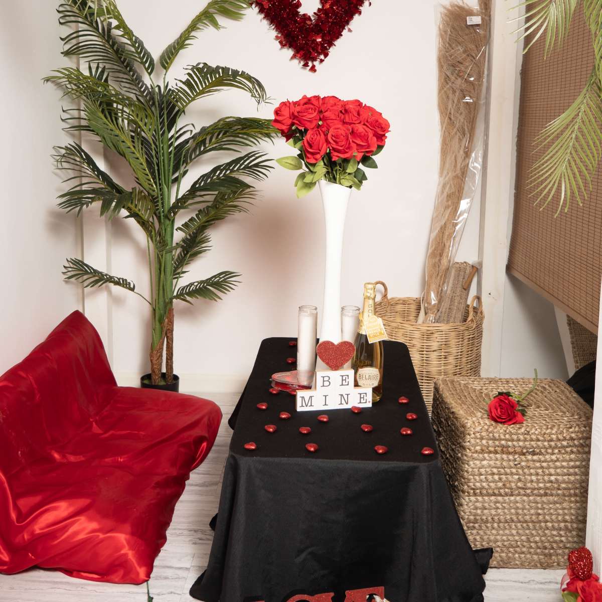 The scene features a table draped in black cloth adorned with candles, rose petals, and a bottle of champagne, set against a backdrop of decorative plants and a heart-shaped wreath.