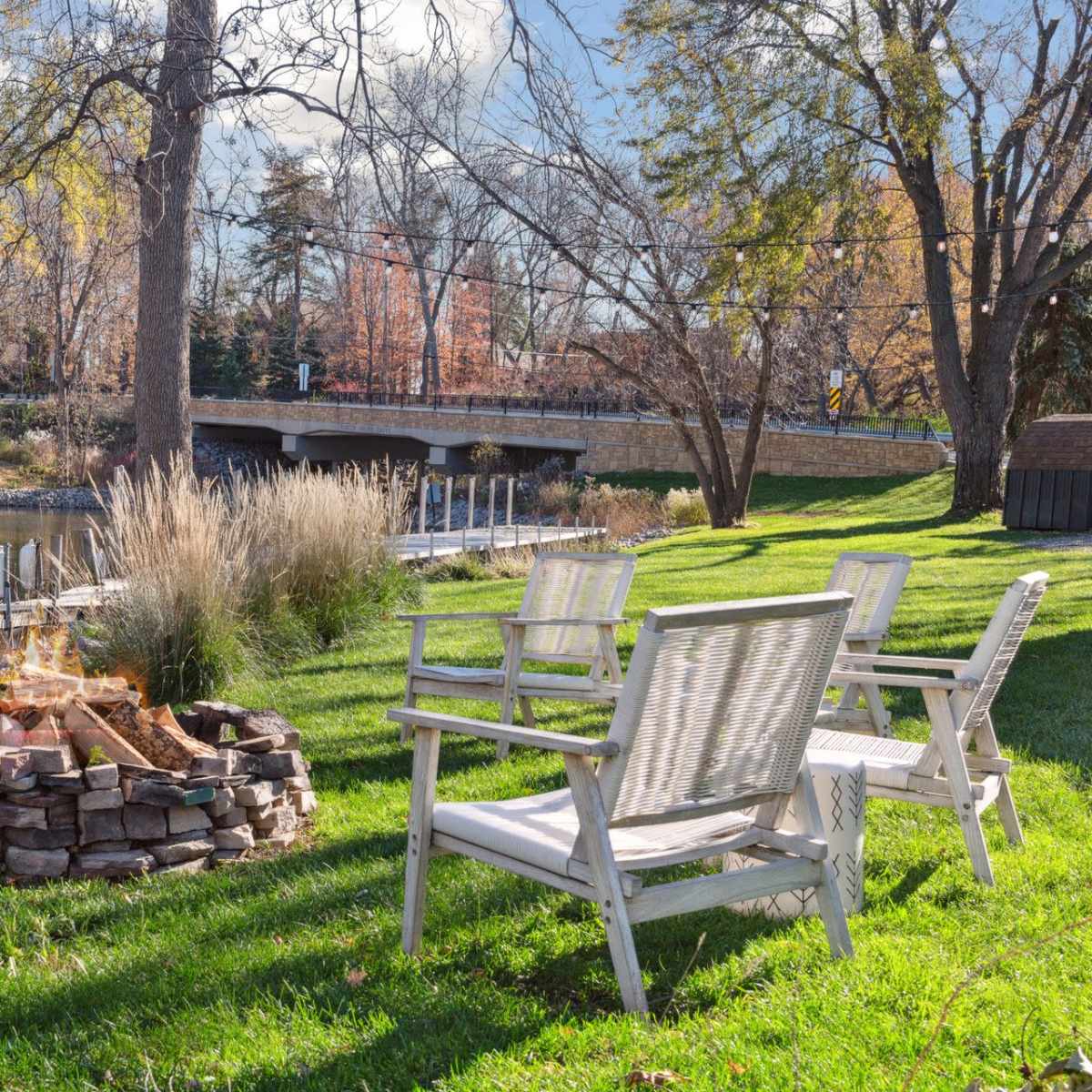 A fire pit surrounded by wooden chairs is situated on a grassy area near a calm pond and a bridge in the background.