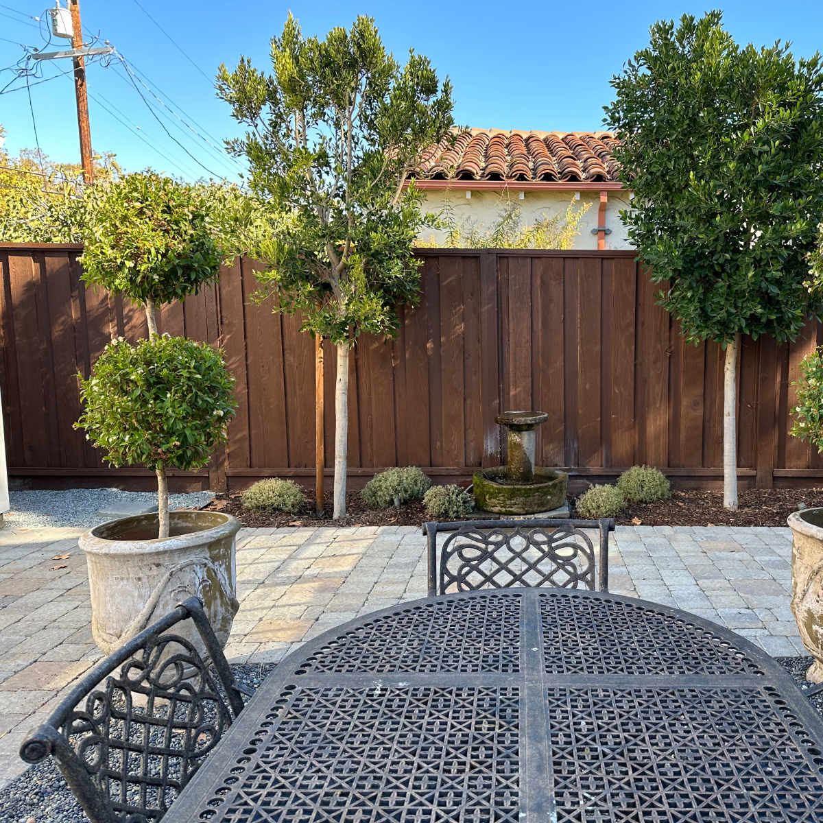 A patio scene features a wrought iron table surrounded by chairs, with two potted topiary trees and a fountain against a wooden fence.