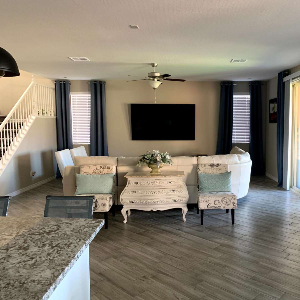 A living room features a white sectional sofa, a mounted TV, a coffee table, and large windows with blue curtains.