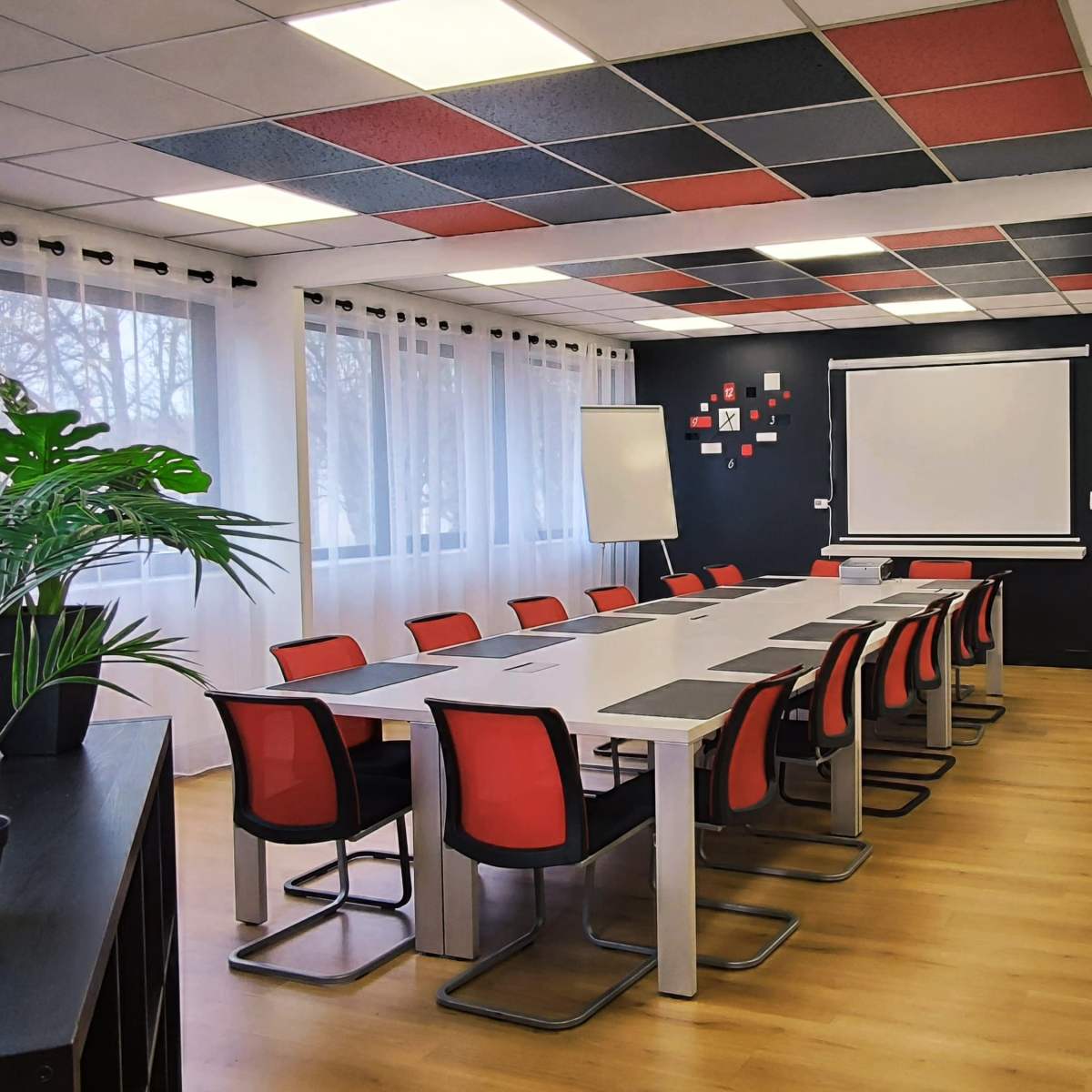 A modern conference room with a long table surrounded by red and gray chairs, a projector screen on the wall, and large windows letting in natural light.