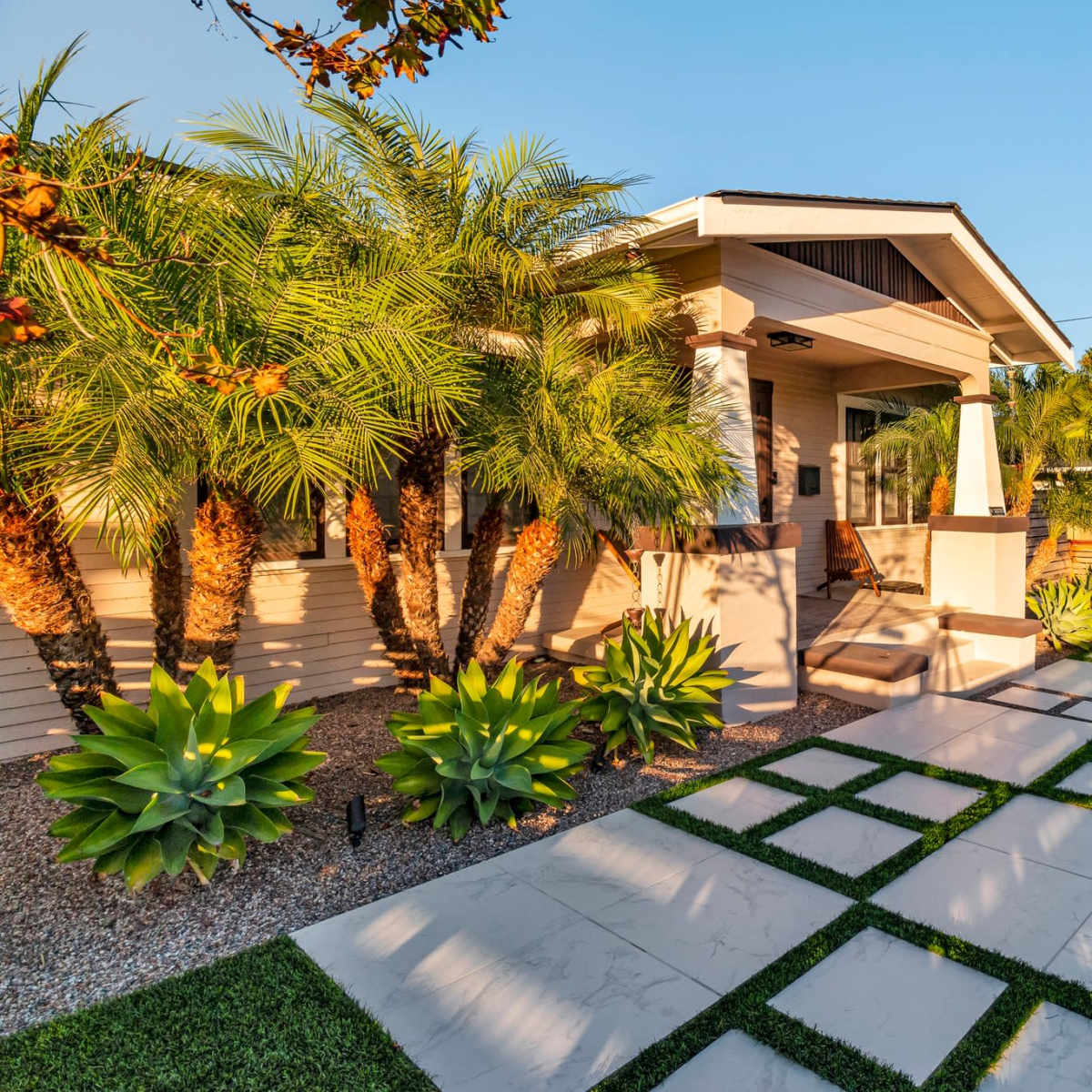 A pathway of large stone tiles leads to a house surrounded by palm trees and succulents in a landscaped yard.