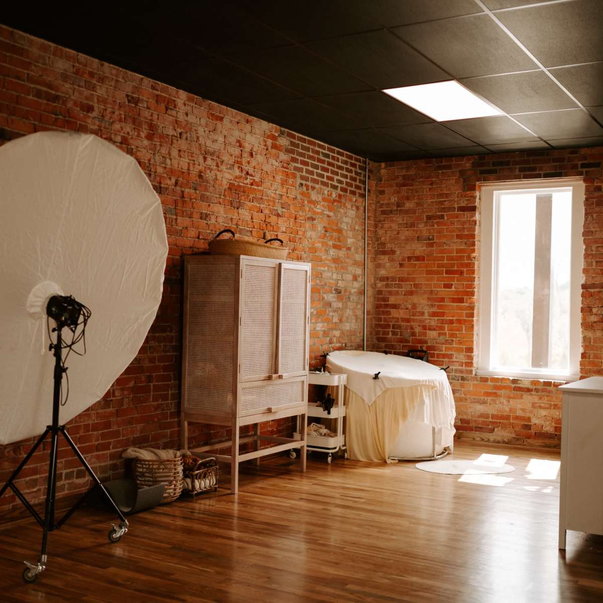 A studio space with exposed brick walls, featuring a large light reflector on a stand, a wooden cabinet, and a massage table beside a window.