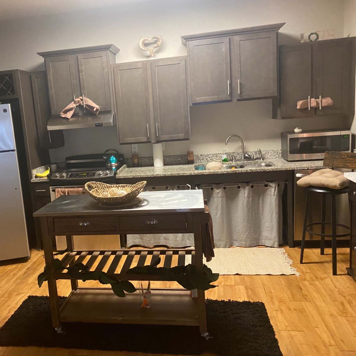 A kitchen with dark cabinets, a silver refrigerator, a wooden island with a decorative bowl, and a black rug on a wood floor.