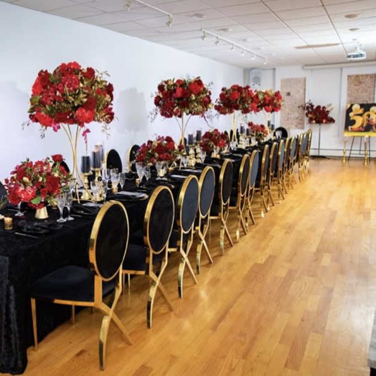A long banquet table is set with black tablecloths and elegant floral arrangements in red, surrounded by gold-accented chairs.