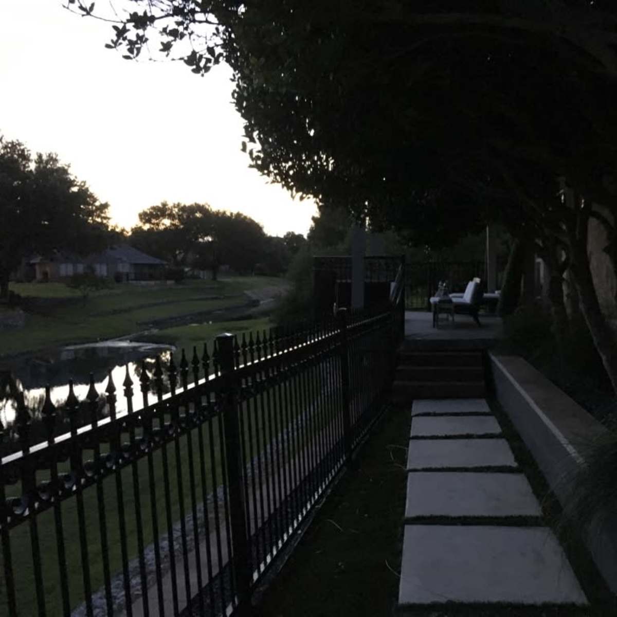A pathway of stone slabs leads alongside a fence, with a view of a calm body of water and trees at dusk.