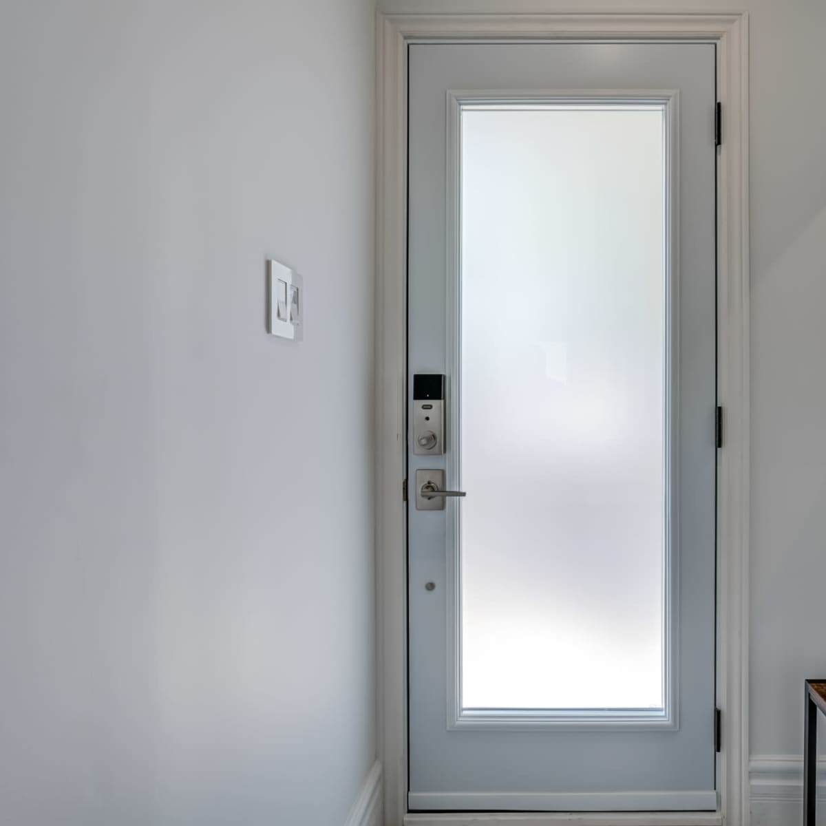 A modern entrance area with a frosted glass door, a wall-mounted mirror, and a wooden bench against a white wall.
