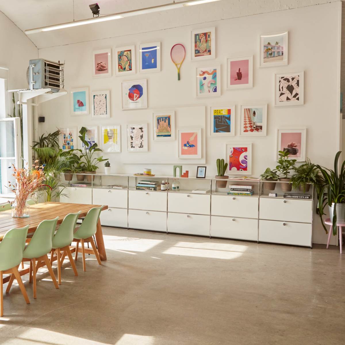A spacious room features a large wooden table with green chairs, surrounded by plants and a wall adorned with various framed artworks.
