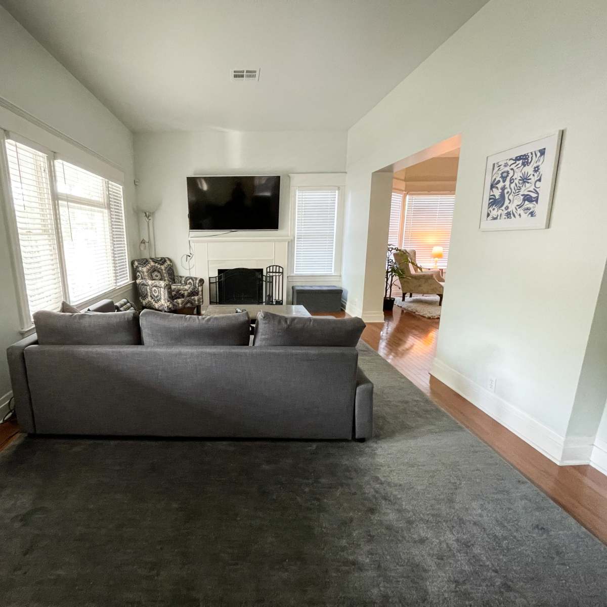 A living room with a gray sofa facing a wall-mounted television, situated near windows with white blinds.