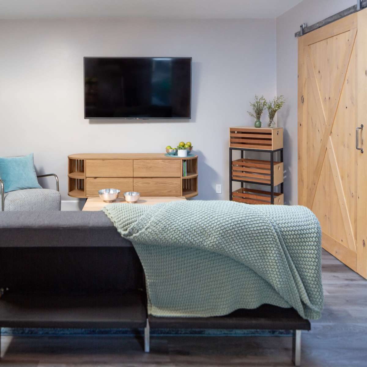 A modern living room features a wall-mounted TV, a wooden media console, a gray sofa with a knitted throw, and natural light coming from a door leading outside.