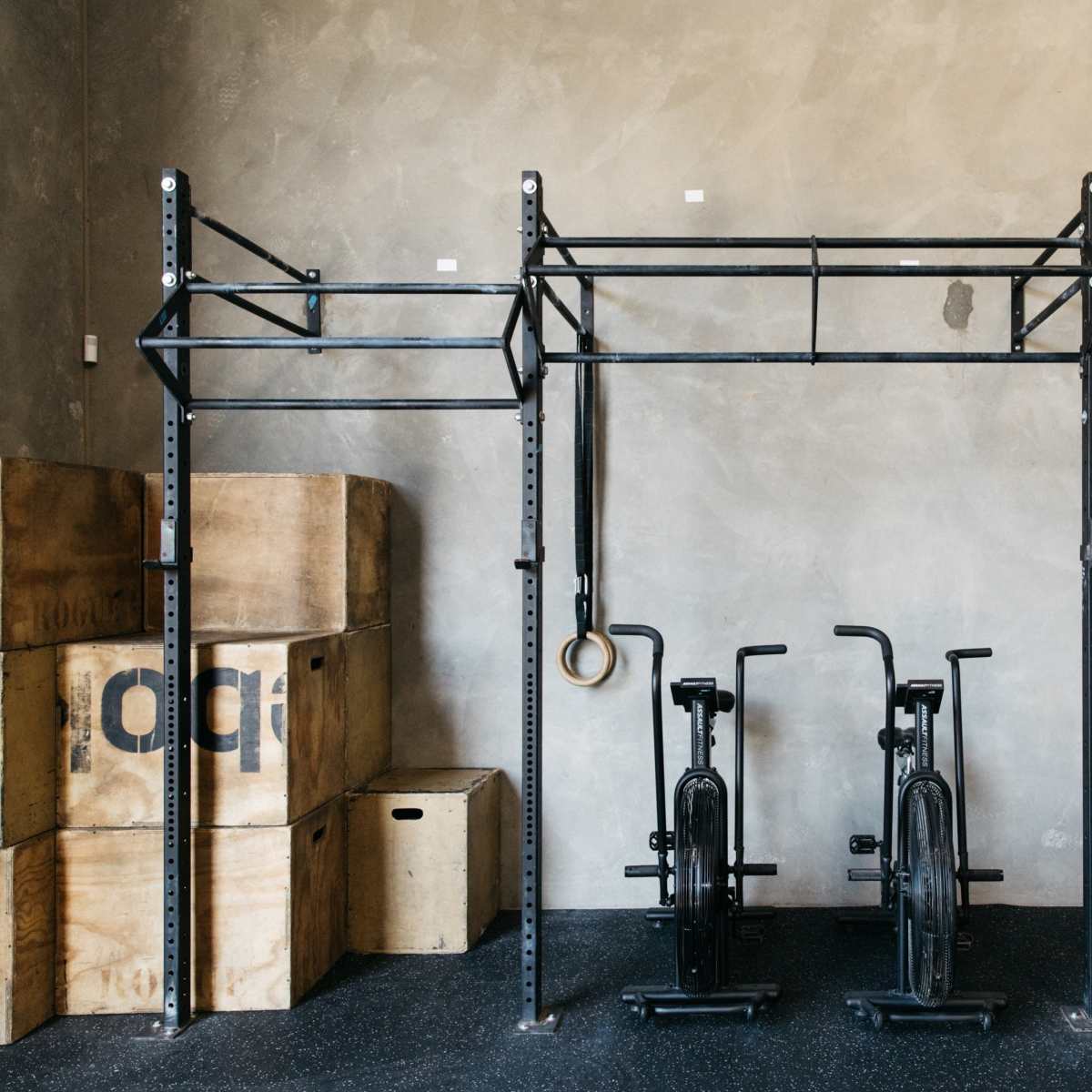 A gym setup with a metal frame for pull-ups, rings hanging from it, and two exercise bikes next to wooden boxes against a grey wall.