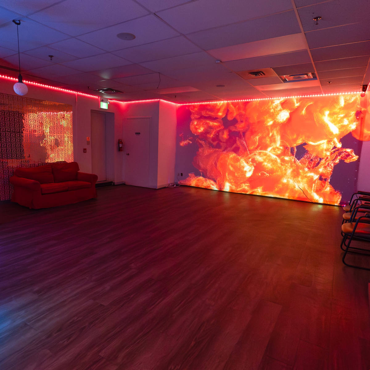 The room features a vibrant wall display of fiery colors, illuminated by neon lights and furnished with a red sofa and chairs.