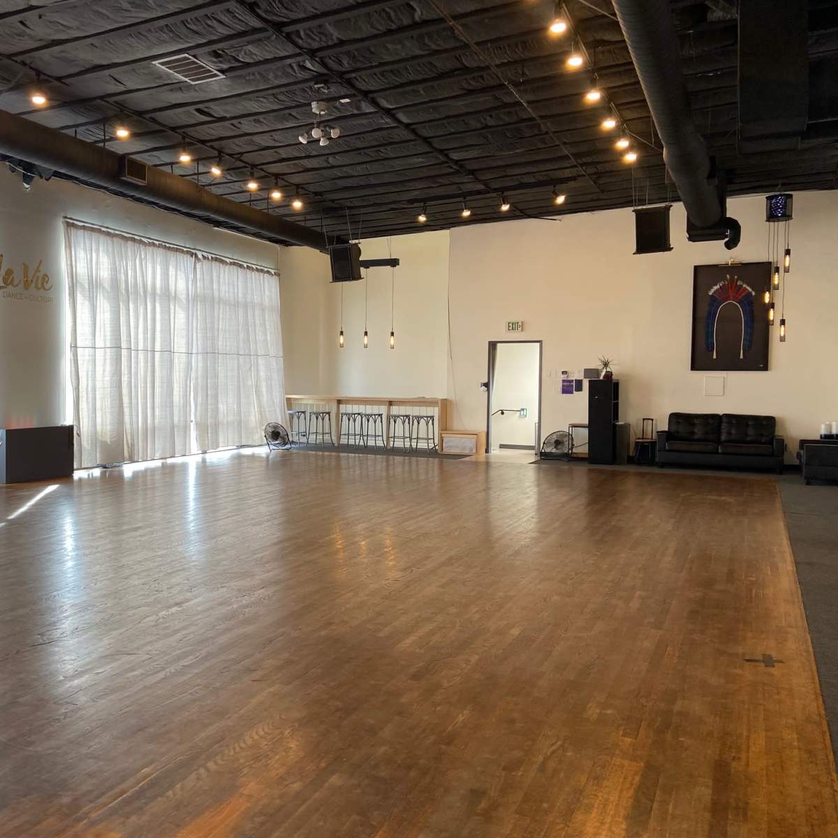 A spacious dance studio with wooden flooring, large windows covered by sheer curtains, and minimalist furnishings including a couch and high tables.