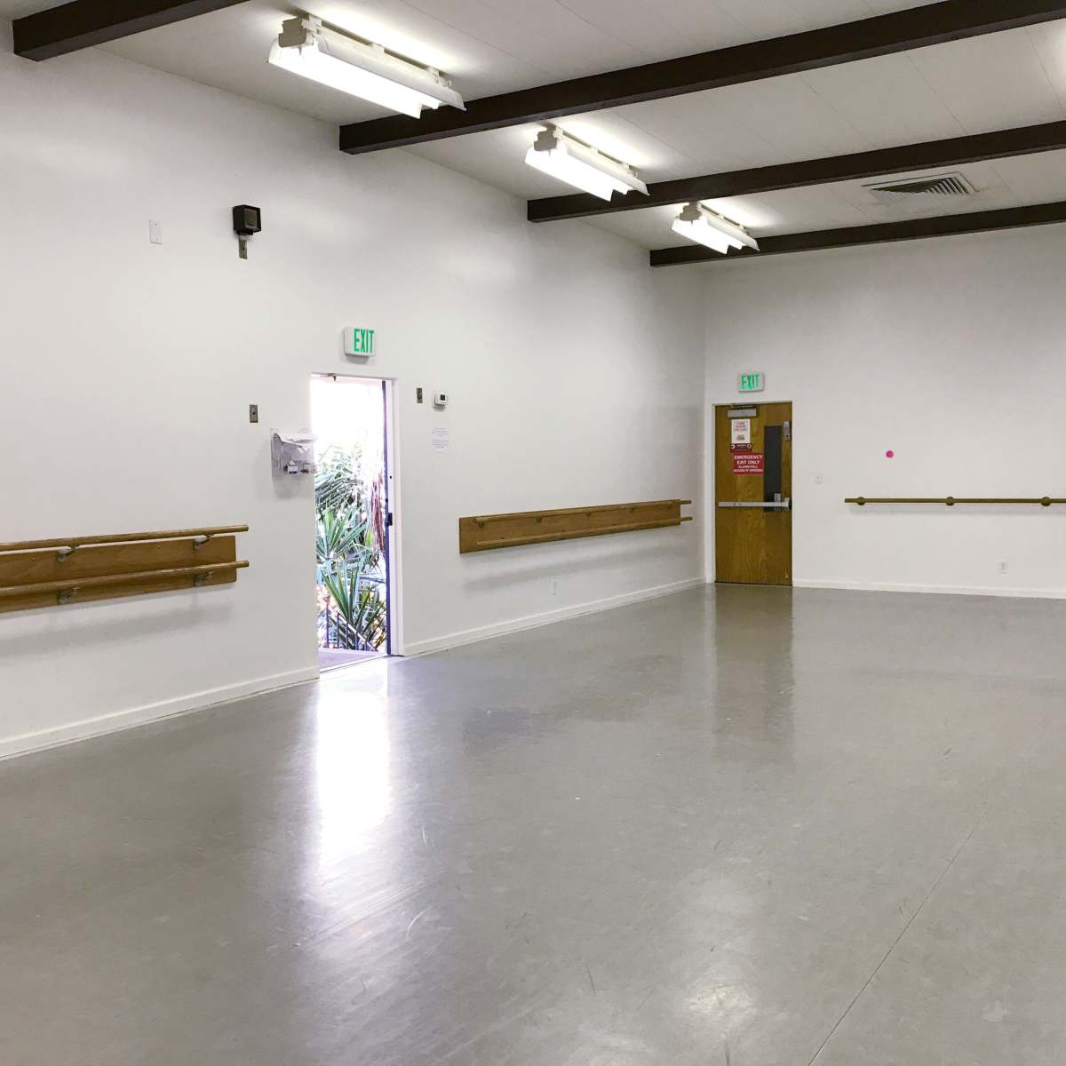 An empty dance studio features mirrored walls, wooden ballet bars, and a door opening to an outdoor area.