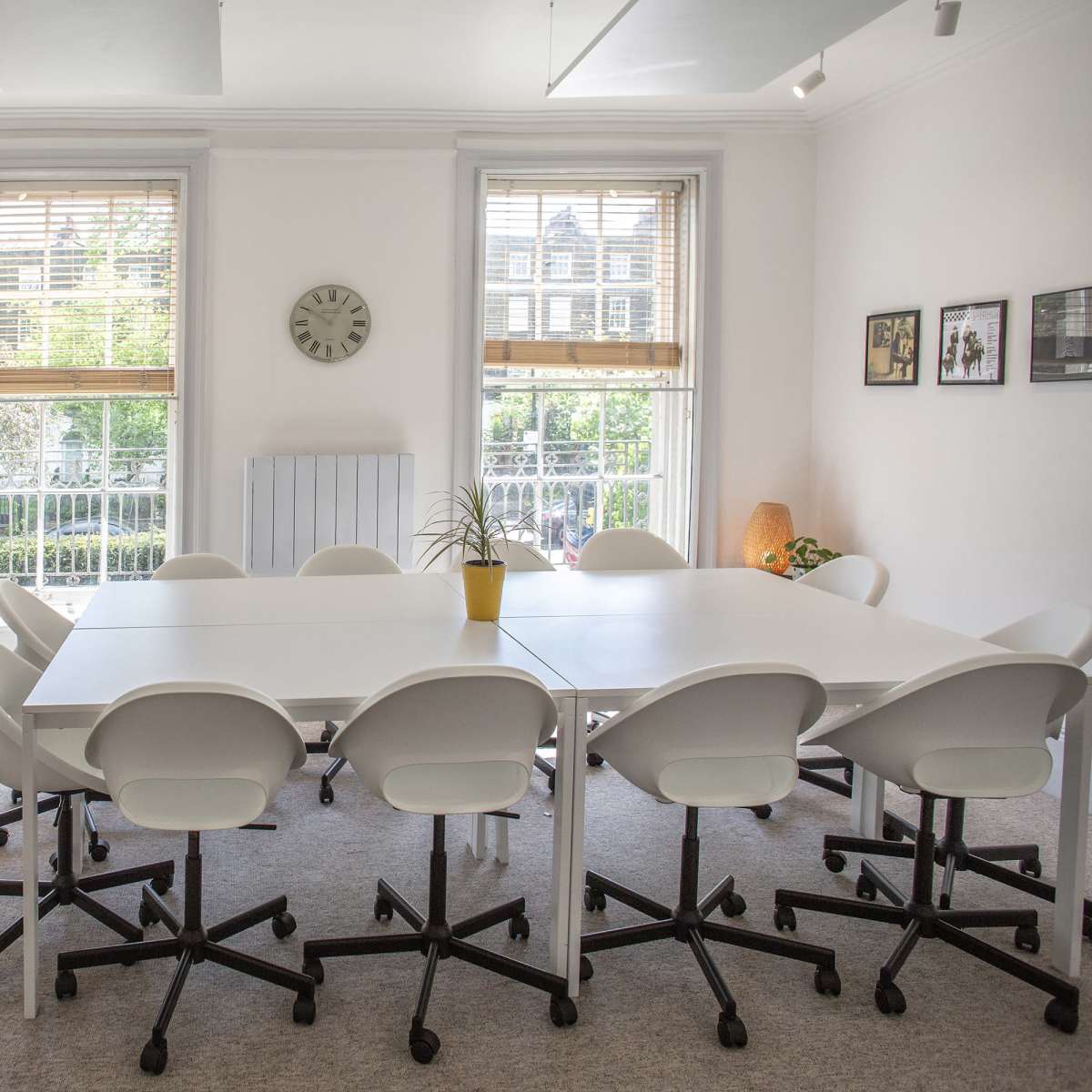 A bright meeting room features a large white table surrounded by chairs, with windows overlooking greenery and a clock on the wall.