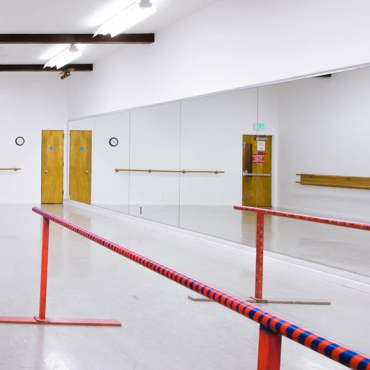 A bright dance studio features a mirrored wall, wooden barre, and double doors with exit signs.