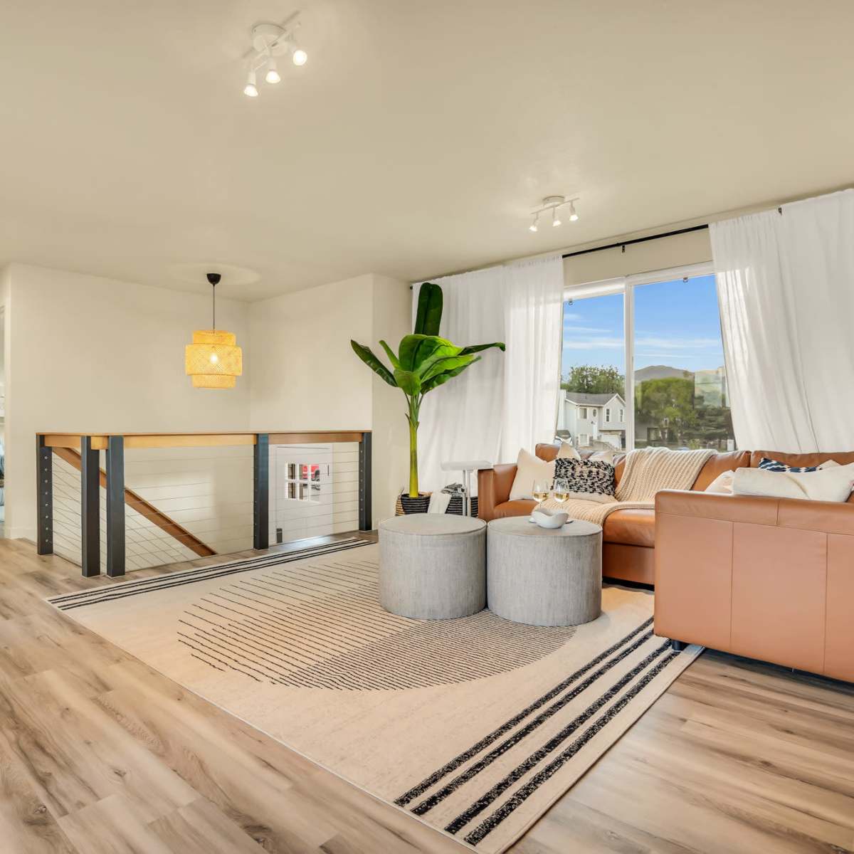 A cozy living room features a brown leather sofa, a large window with white curtains, and two round side tables on a patterned rug.