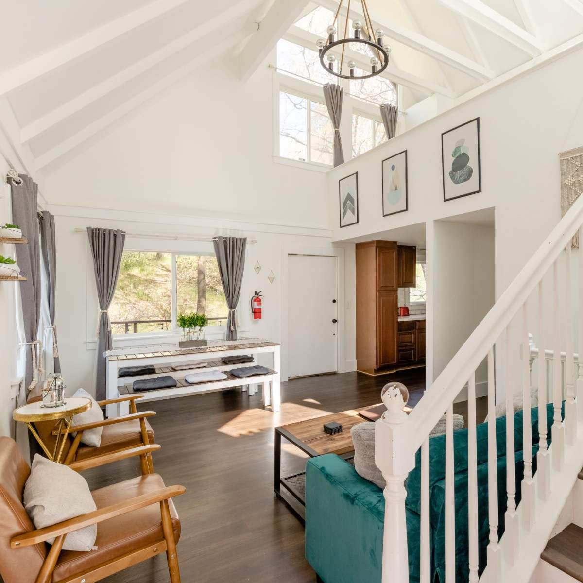 A bright, airy living room featuring a staircase, a teal couch, wooden chairs, and large windows with natural light.
