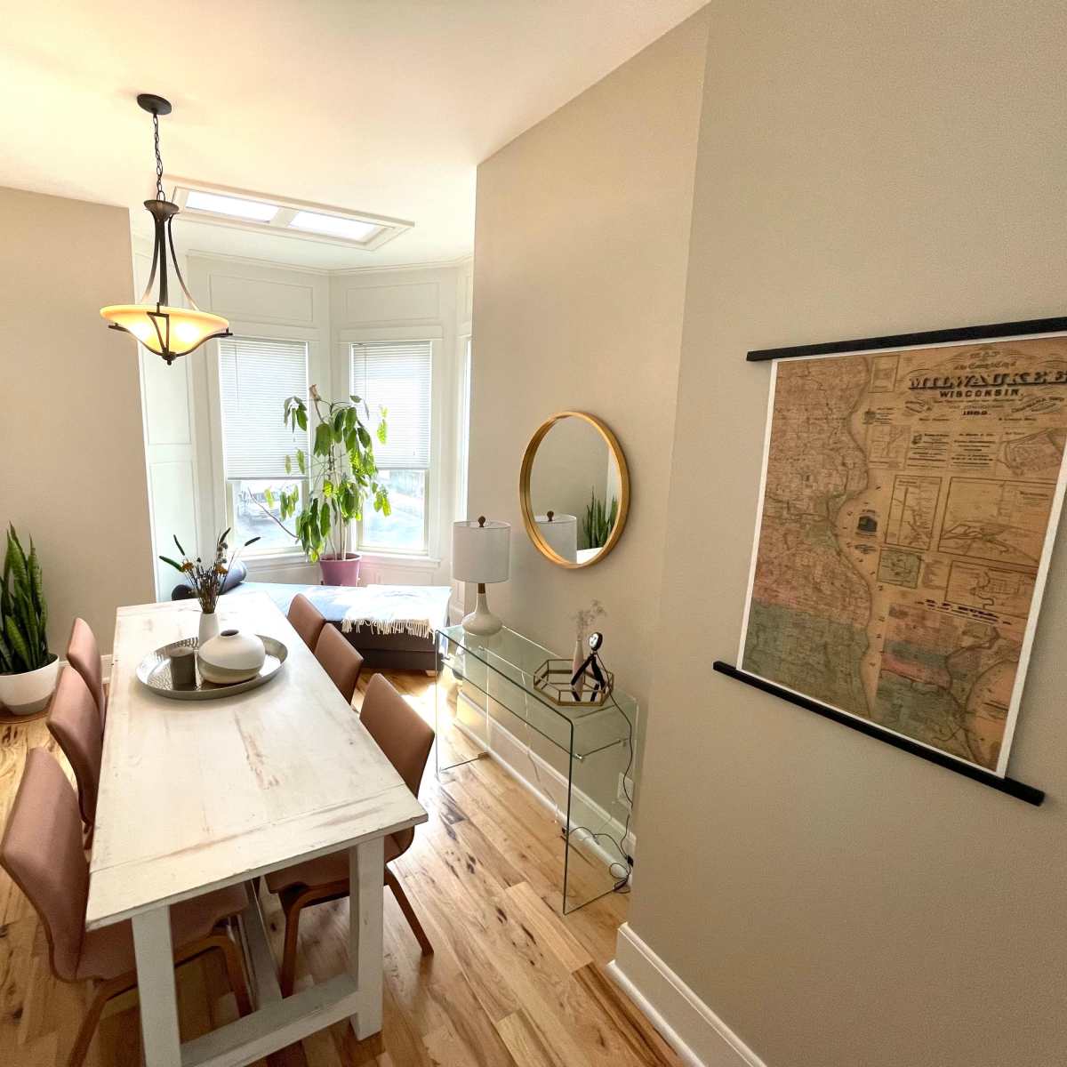 A dining area with a wooden table surrounded by four chairs, a large window, a potted plant, and framed decor on the walls.