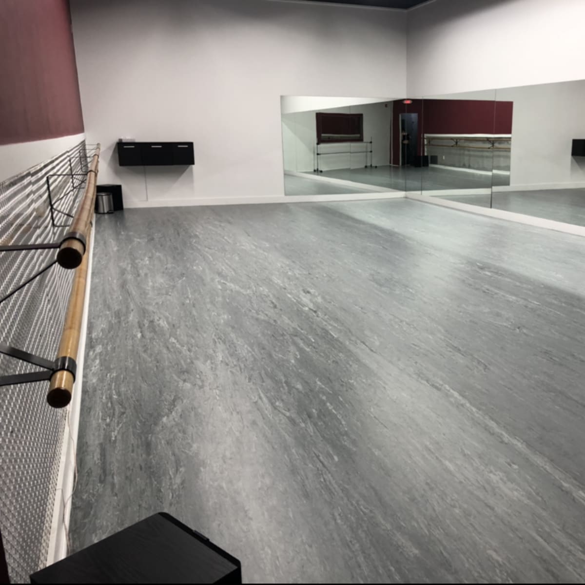A spacious dance studio with a mirrored wall, wooden ballet bar, and gray flooring.